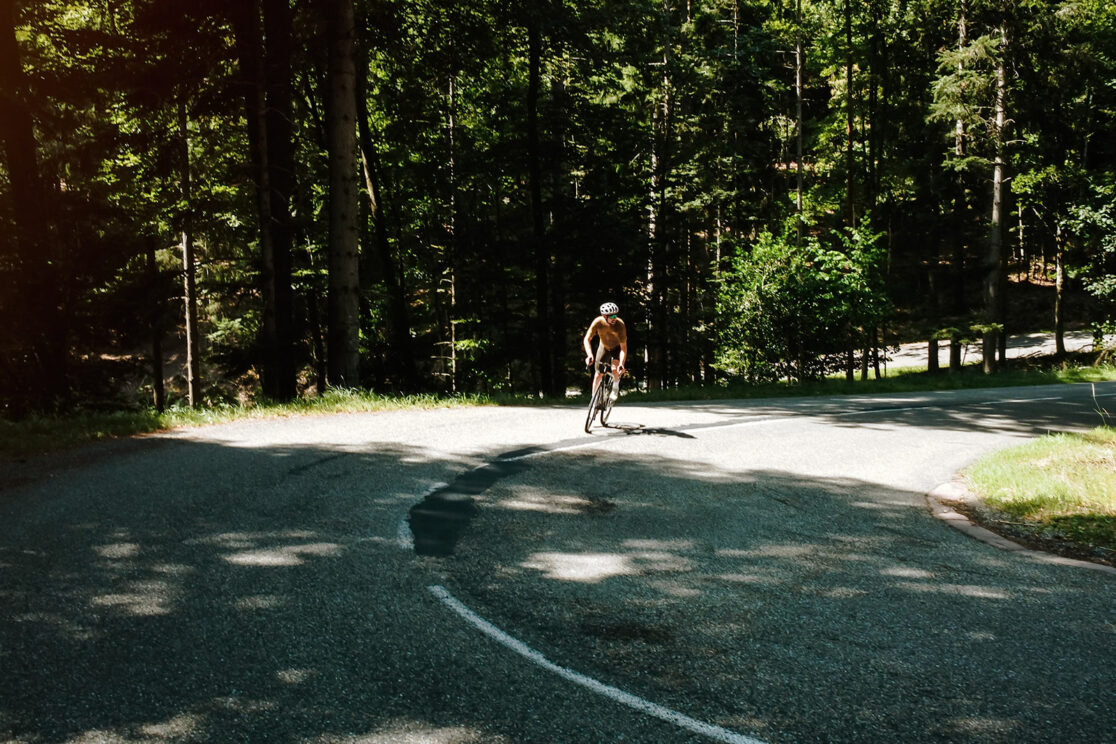 Rennradfahrer biegt im Schatten eines Waldes um die Kurve, Sonnenflecken auf der Fahrbahn, Rennradfotografie von Sebastian Weindel