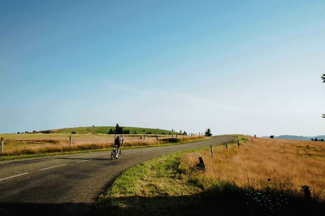 Radfahrer auf schmaler Straße durch weite Graslandschaft unter blauem Himmel, Rennradfotografie von Sebastian Weindel