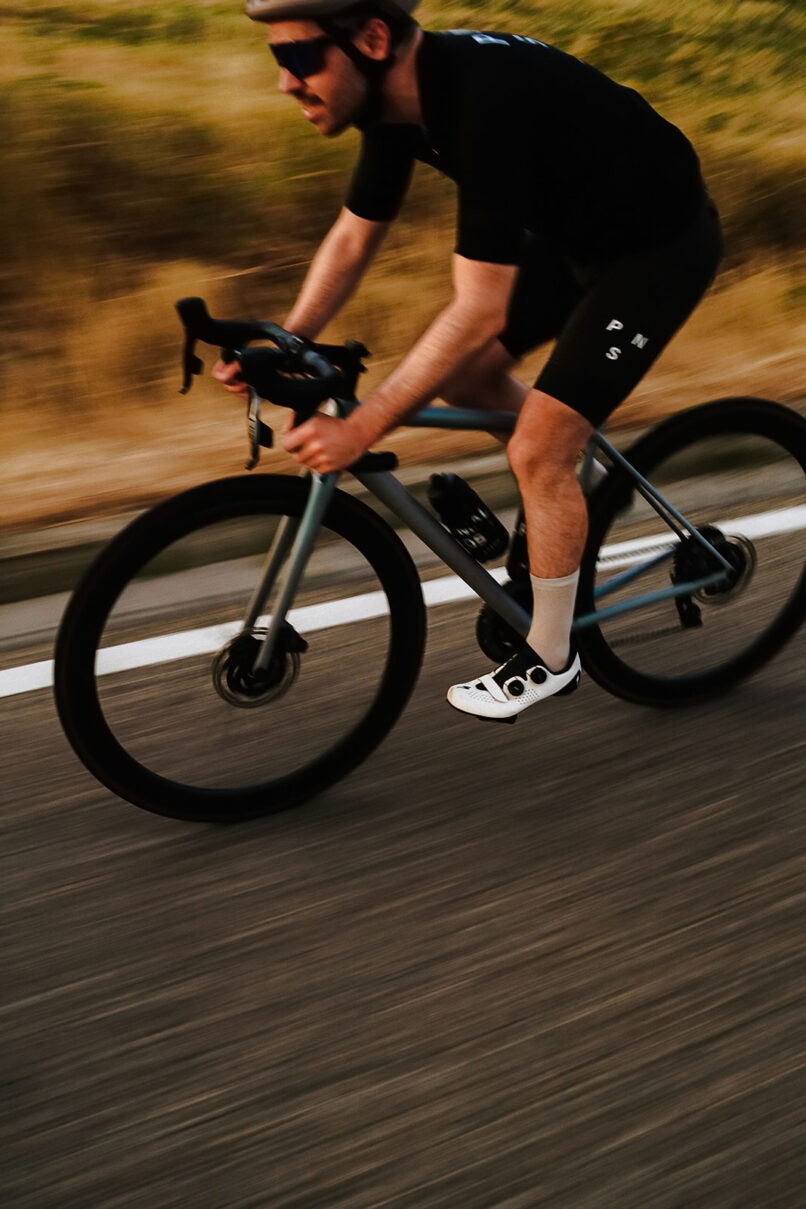 Rennradfahrer in schwarzem Trikot und weißen Schuhen, Bewegungsunschärfe auf Asphalt, Rennradfotografie von Sebastian Weindel