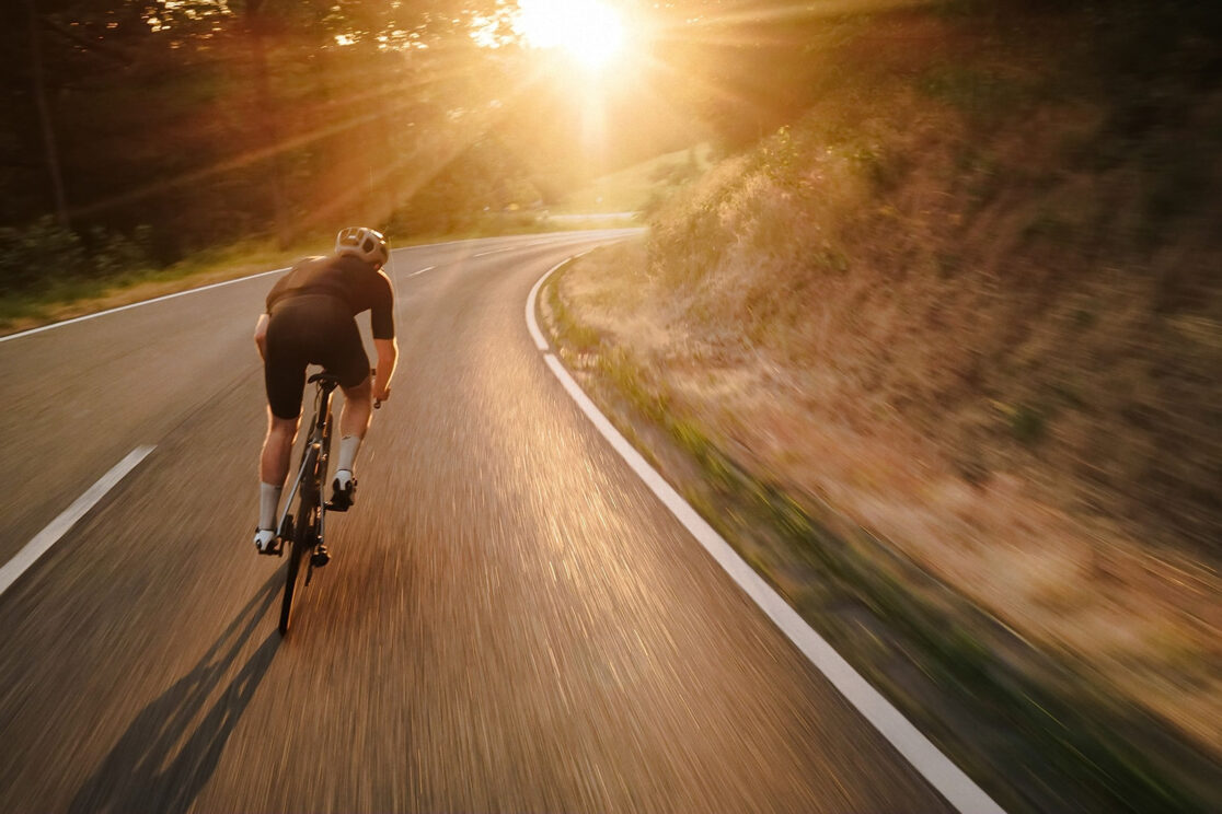 Radfahrer von hinten in einer Kurve, tiefe Sonne über der Straße, Bewegungsunschärfe, Rennradfotografie von Sebastian Weindel