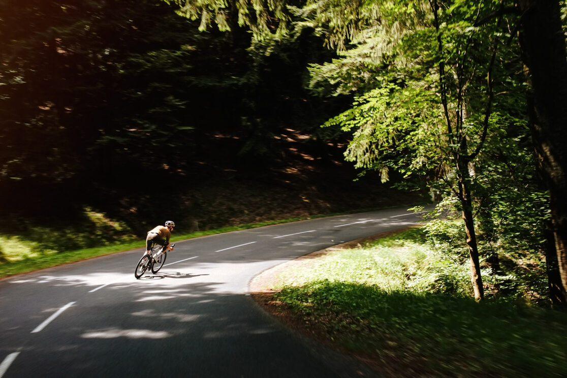 Radfahrer in gelbem Trikot in einer Kurve unter Laubbäumen, Schatten auf der Straße, Rennradfotografie von Sebastian Weindel