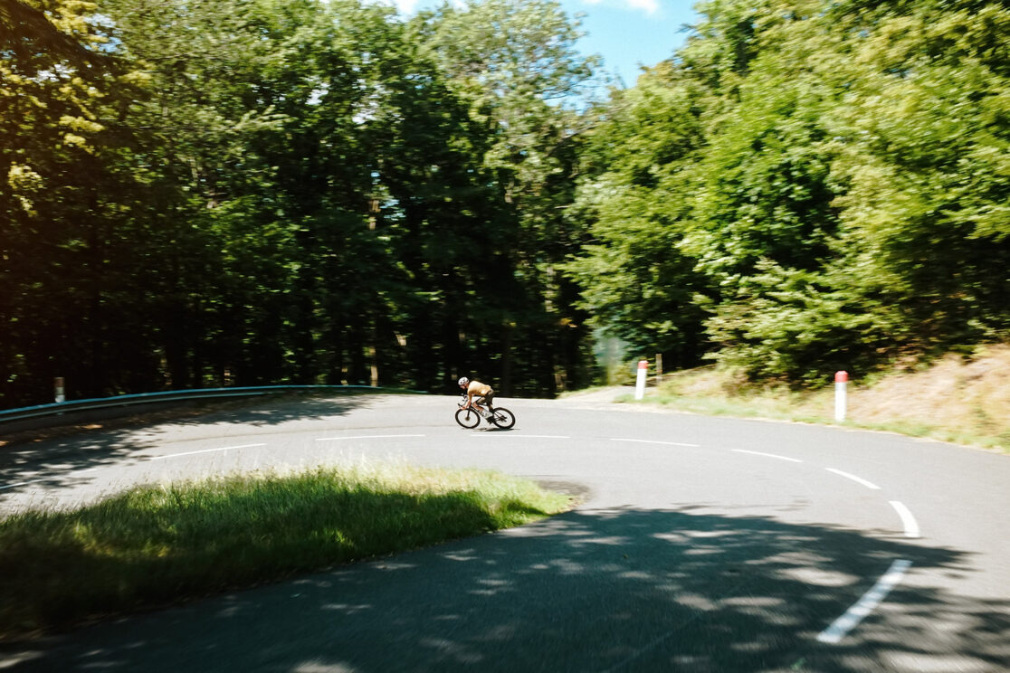 Winziger Radfahrer im Licht am Ende einer dunklen Waldstraße, Leitplanke rechts, Rennradfotografie von Sebastian Weindel