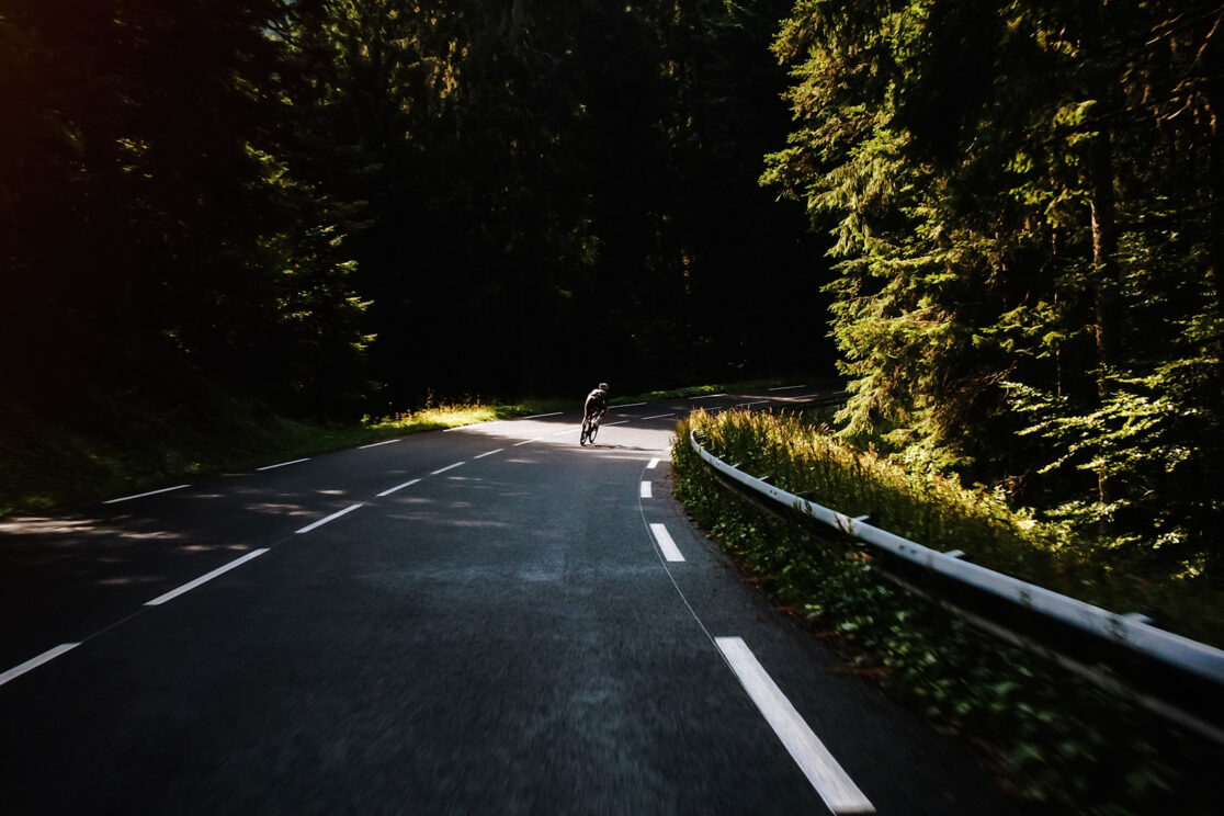 Radfahrer in dunklem Trikot fährt eine Kurve entlang der Leitplanke, grüne Bäume, Sonne, Rennradfotografie von Sebastian Weindel