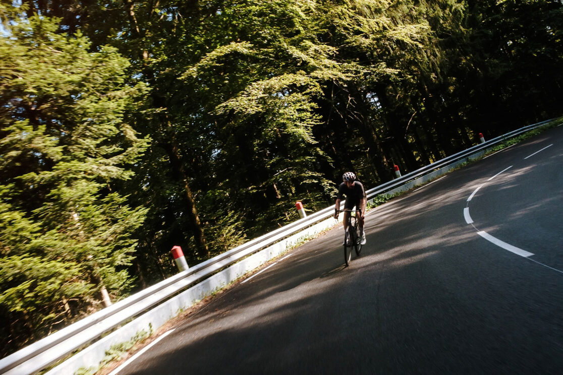 Radfahrer kommt auf die Kamera zu, Kurve an der Leitplanke durch dichten Wald, Rennradfotografie von Sebastian Weindel