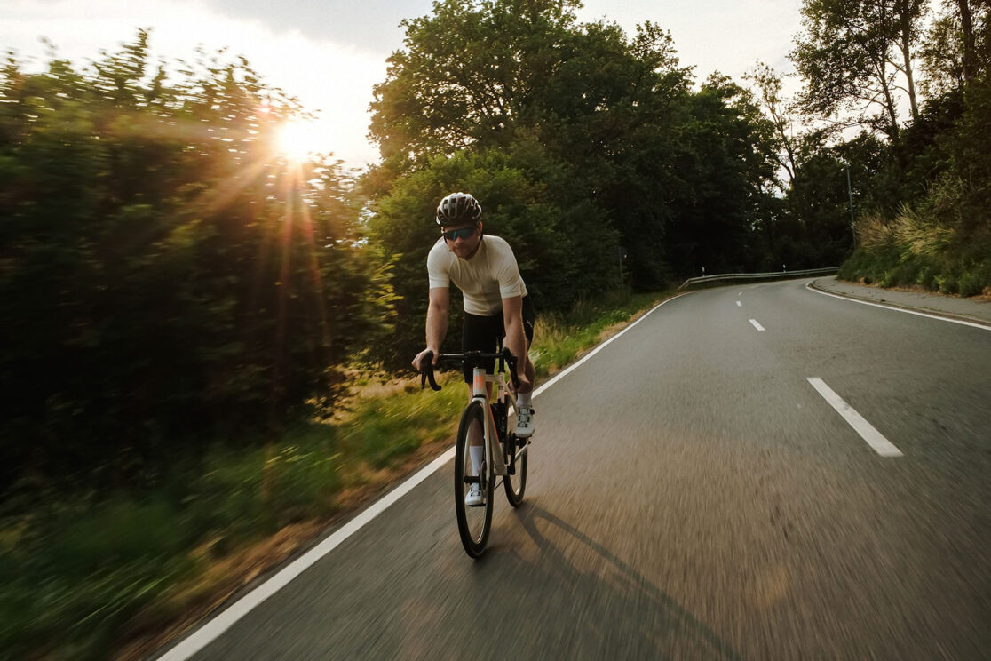 Radfahrer im weißen Trikot auf kurviger Landstraße bei Sonnenuntergang, Rennradfotografie von Sebastian Weindel