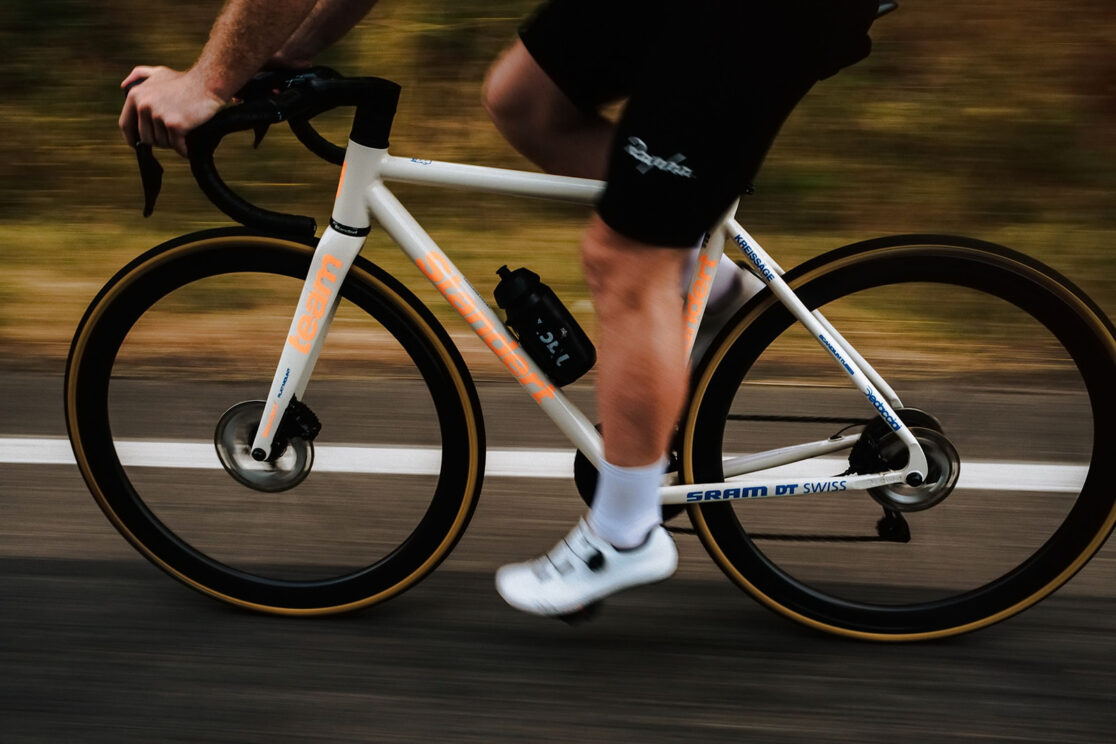 Weißes Rennrad mit orangefarbener Schrift in voller Fahrt, Mitzieher mit verwischtem Hintergrund, Rennradfotografie von Sebastian Weindel