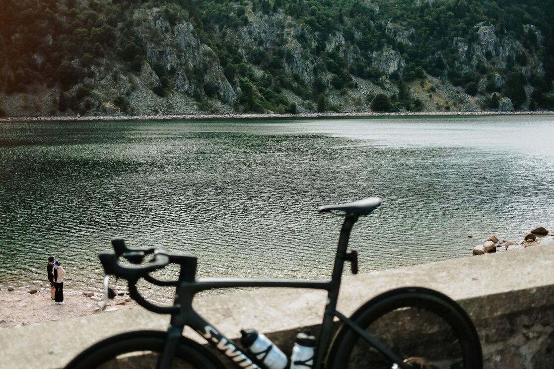 Rennrad an einer Mauer vor einem Bergsee mit Felshängen, zwei Menschen am Ufer, Rennradfotografie von Sebastian Weindel